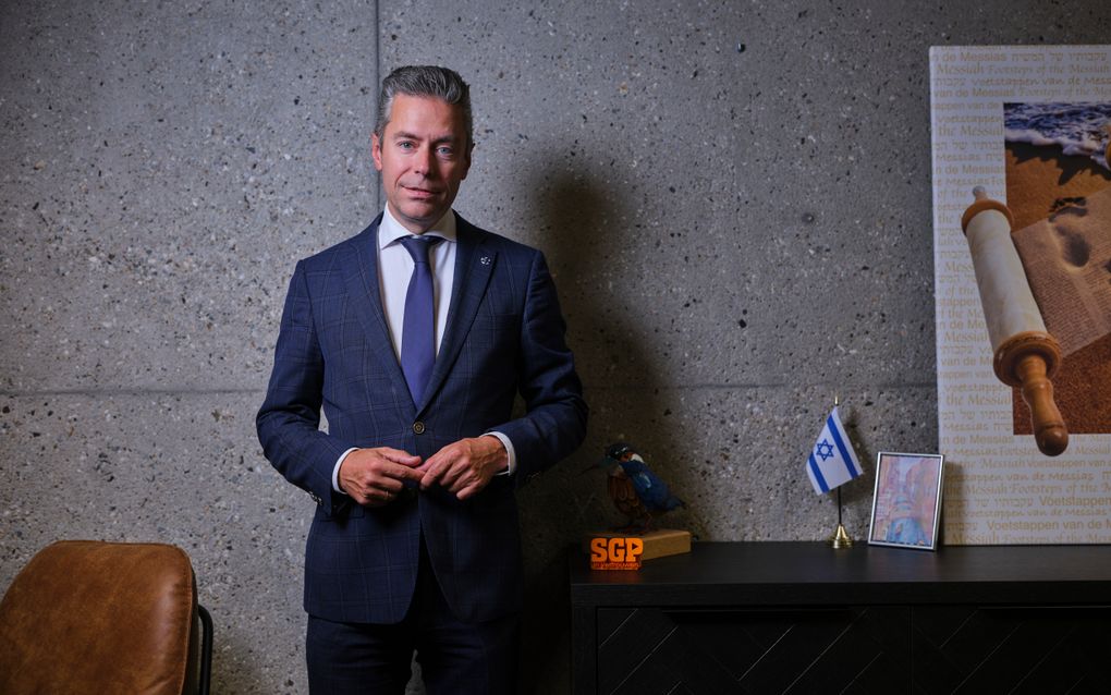 Man in pak staat voor een betonnen muur. Naast hem staan onder meer een SGP-logo en een Israëlische vlag.