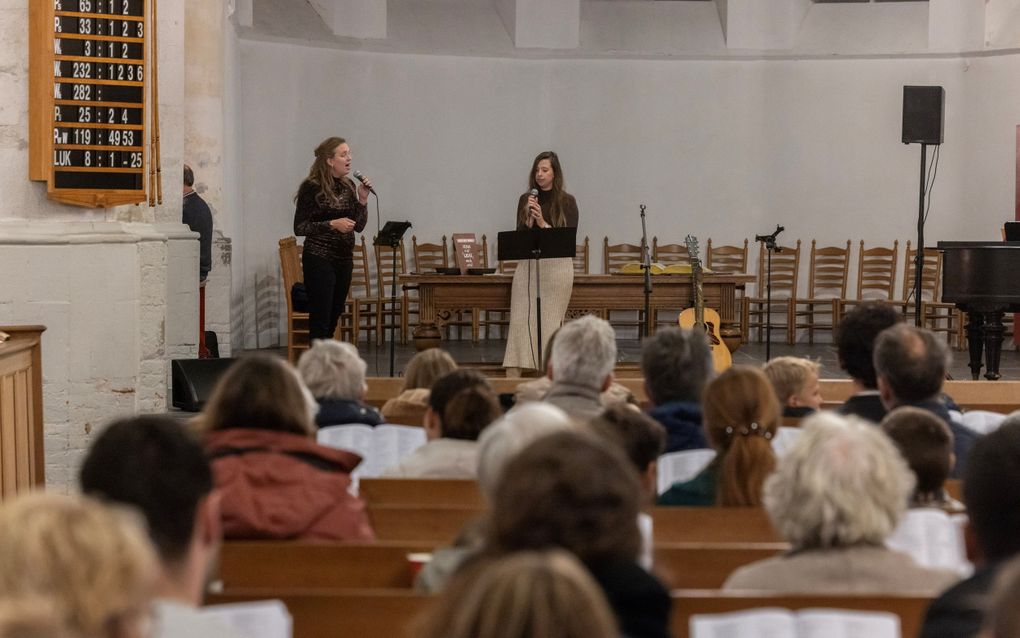 Twee vrouwen staan met een microfoon in de hand, in de banken zitten enkele tientallen mensen.