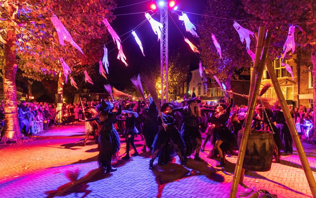 Een groep vrouwen in zwarte kleding en met bezems op een plein. In de lucht hangen witte spookjes.