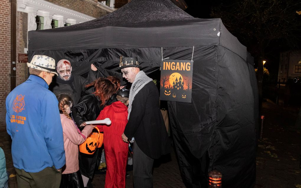 Twee mannen met schmink op hun gezicht staan voor een zwarte tent. Een paar kinderen met oranje tasjes in de vorm van een pompoen lopen naar binnen.