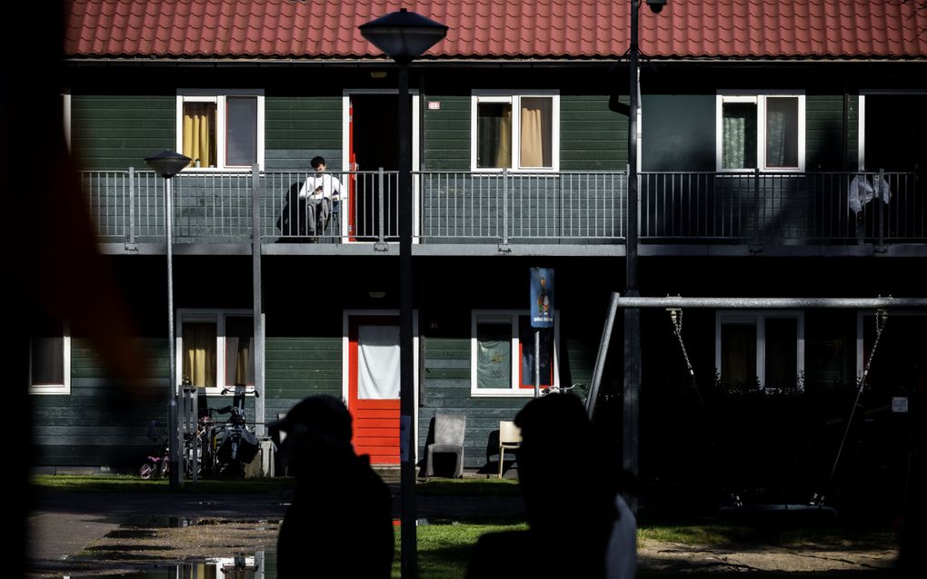 Twee personen lopen langs een gebouw met twee verdiepingen. Op de eerste verdieping zit een asielzoeker op de galerij.