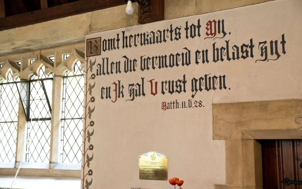 Binnenkant van een kerkgebouw met kleine gotische ramen en laag houten plafond met uitzicht op een gepleisterde muur met daarop Mattheüs 11:28 geschreven in zwarte en rode gotische letters.