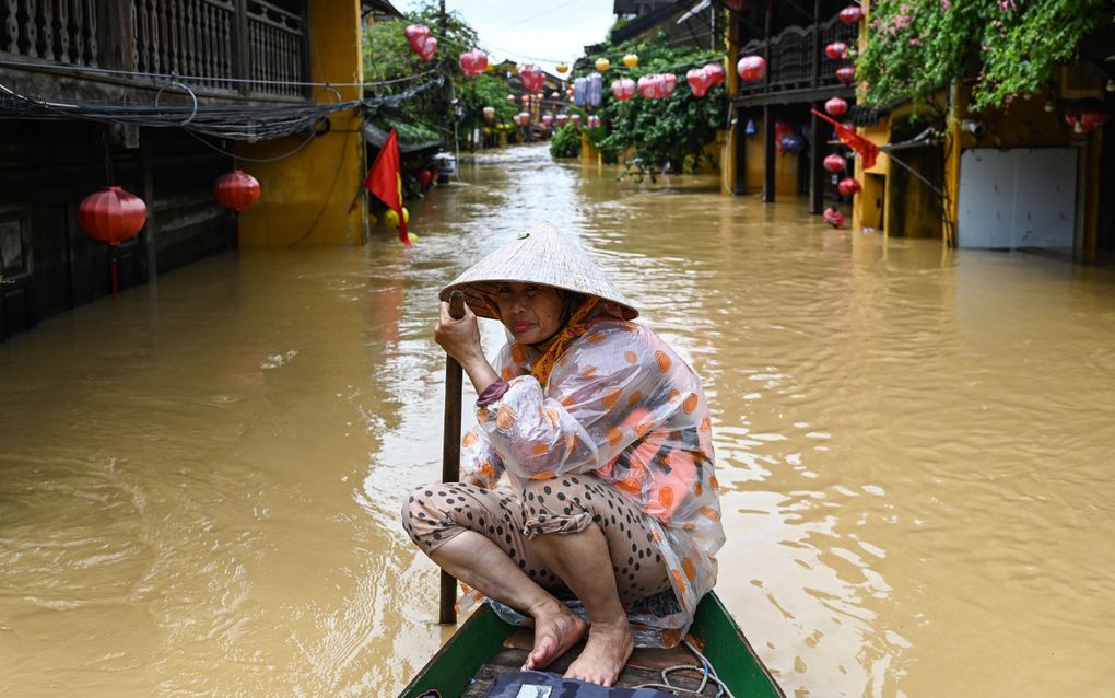 Een vrouw zit op de punt van een boot met een roeiboom in de hand. Ze draagt een vietnamese hoed. Aan weerszijden van het bruine water zijn huizen te zien.