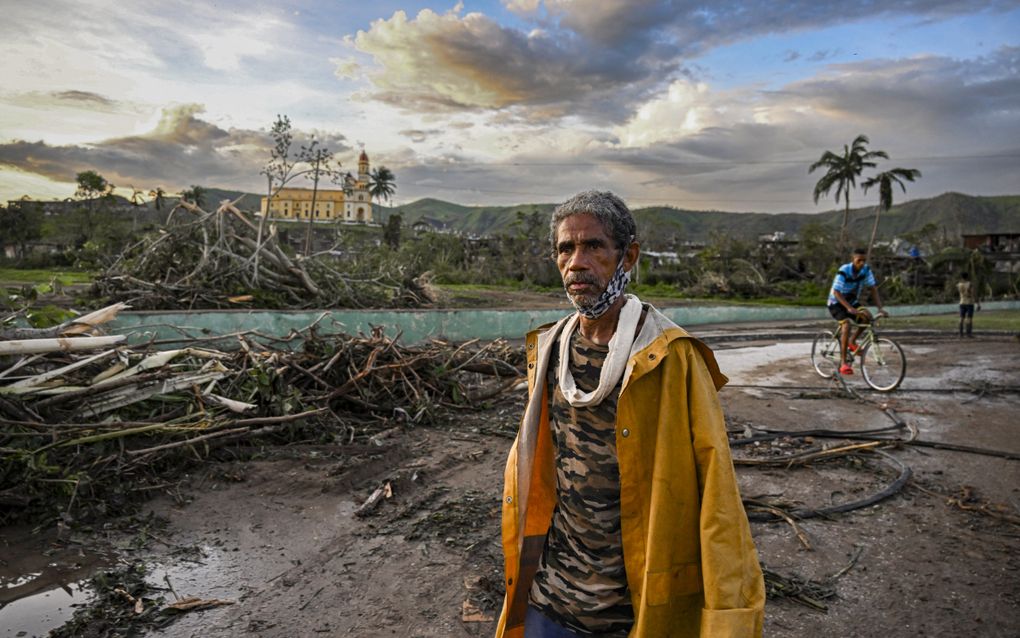 Oudere man in verschoten gele jas en met grijze baard loopt verdwaasd door straten vol puin en hout. op de achtergrond een geel kerkgebouw en bergen.