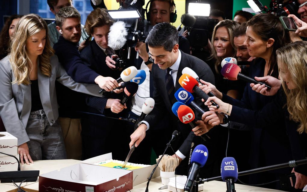 Jonge man in donker pak snijdt een taart aan, terwijl hij omringt wordt door microfoons en camera’s