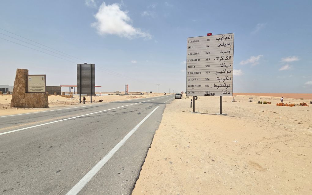 Een geasfalteerde weg loopt door het zand. Aan de rand van de weg staan borden, waarvan een met plaatsnamen en afstanden.