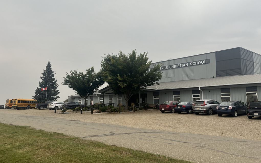 Rechthoekig grijs schoolgebouw met gele schoolbussen en auto’s ervoor.