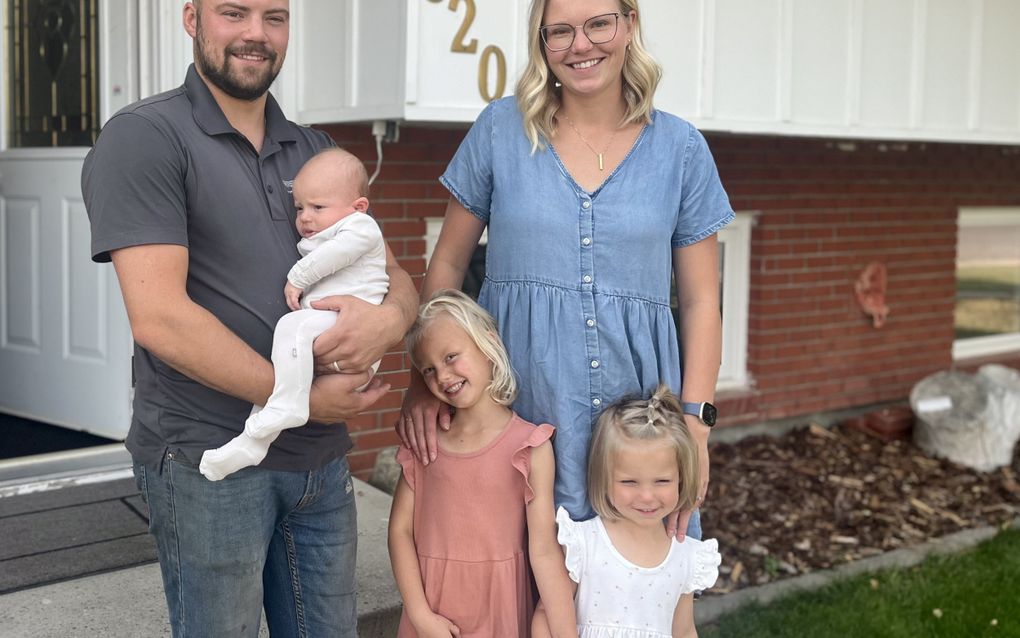 Een jonge vader en moeder met drie kleine kindertjes voor een huis van hout en steen.