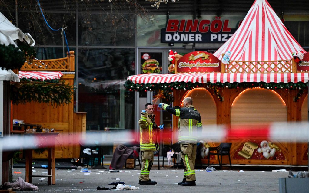 Twee brandweermannen in reflecterende kleding met elkaar in gesprek op de markt, tussen de bruinrode kraampjes.
