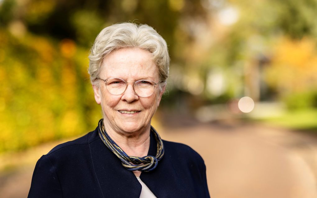 Oude mevrouw met grijs, opgestoken haar, montuurloze bril, glimlach, donker jasje, en blauw met gouden collier.