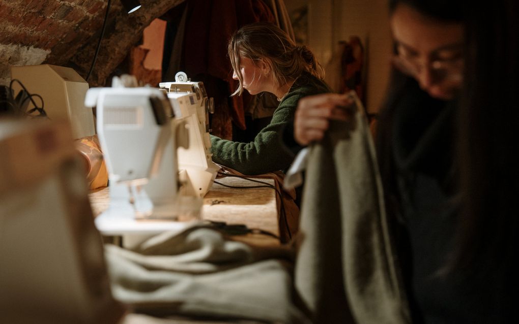 Een blonde vrouw in groene trui zit ingespannen achter de naaimachine. Op de voorgrond is een andere vrouw bezig met een lichtgroen kledingstuk. 