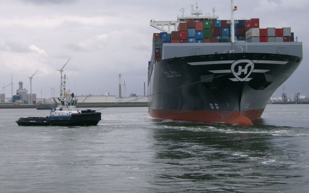 Containerschip in de haven, met heel veel gekleurde containers op het dek. Daarnaast een sleepboot.