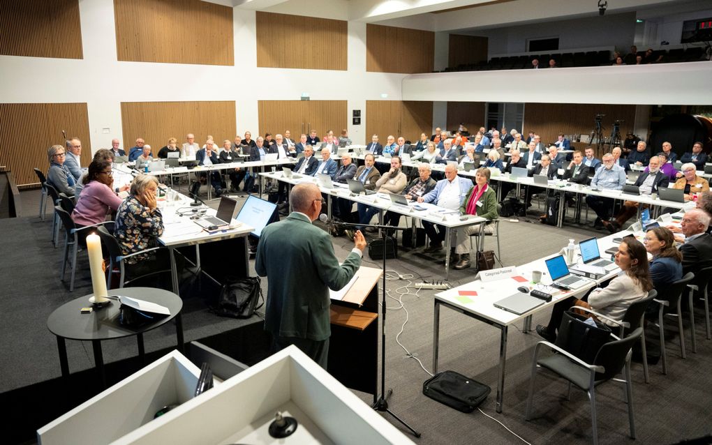Vergaderzaal met tafels waarachter mannen en vrouwen zitten; een man achter een microfoon houdt een toespraak.