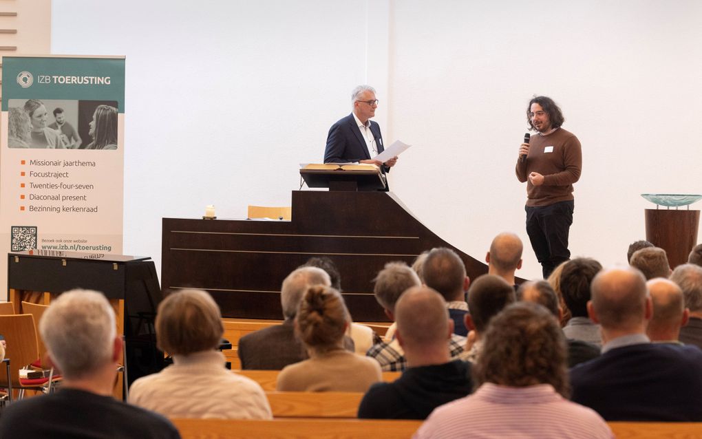 Man met een trui, vrij lang haar, doet zijn verhaal via de microfoon. Links ds. Van der Graaf.