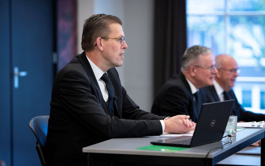 Dominee Mulder, een man in zwart pak, bril op, zit achter een tafel, een laptop opengeklapt.