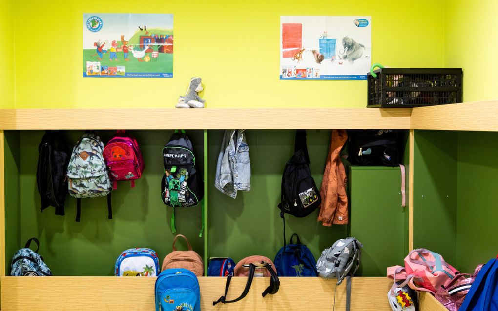 Verschillende kleuren tassen en twee kinderjassen hangen aan kapstok met groene achtergrond. Op de kapstok ligt knuffel en erboven hangen twee posters met dieren.