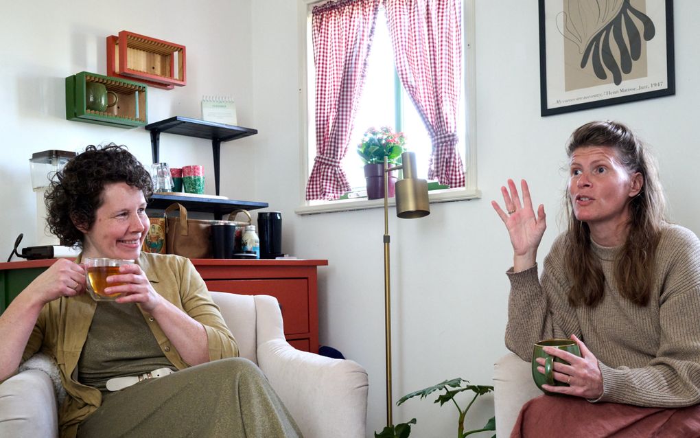 Vrouw met donkere krullen zit links in een witte fauteuil. Ze heeft een kop thee in haar handen. Rechts zit een vrouw met lang blond haar. Ze steekt haar rechterhand op.