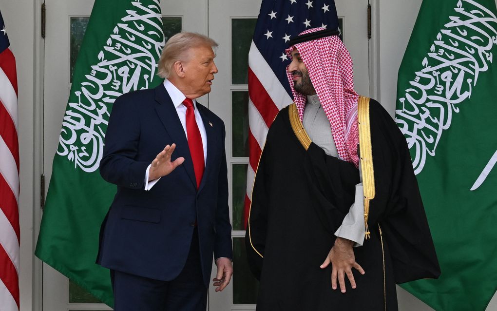 Twee mannen, de een in pak en de ander in zwart gewaad met rood geruite hoofddoek, staan voor een wit gebouw. Achter hen zijn Amerikaanse en Saudische vlaggen te zien 