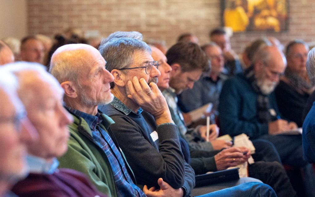 Rijen mensen die zitten te luisteren. Een man maakt aantekeningen. 