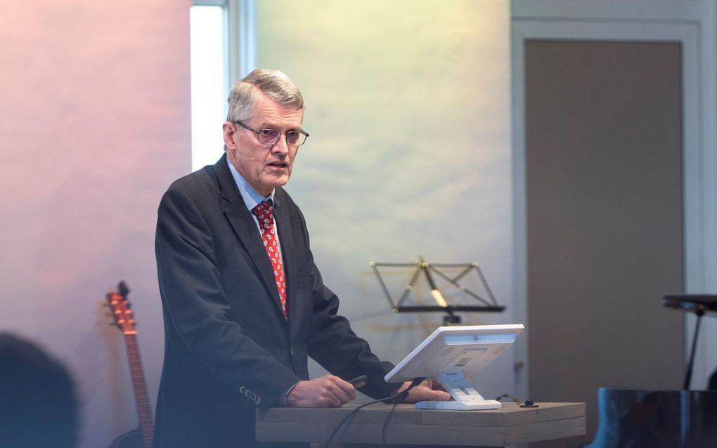 Man met donker pak, grijs haar en bril staat achter een katheder. 