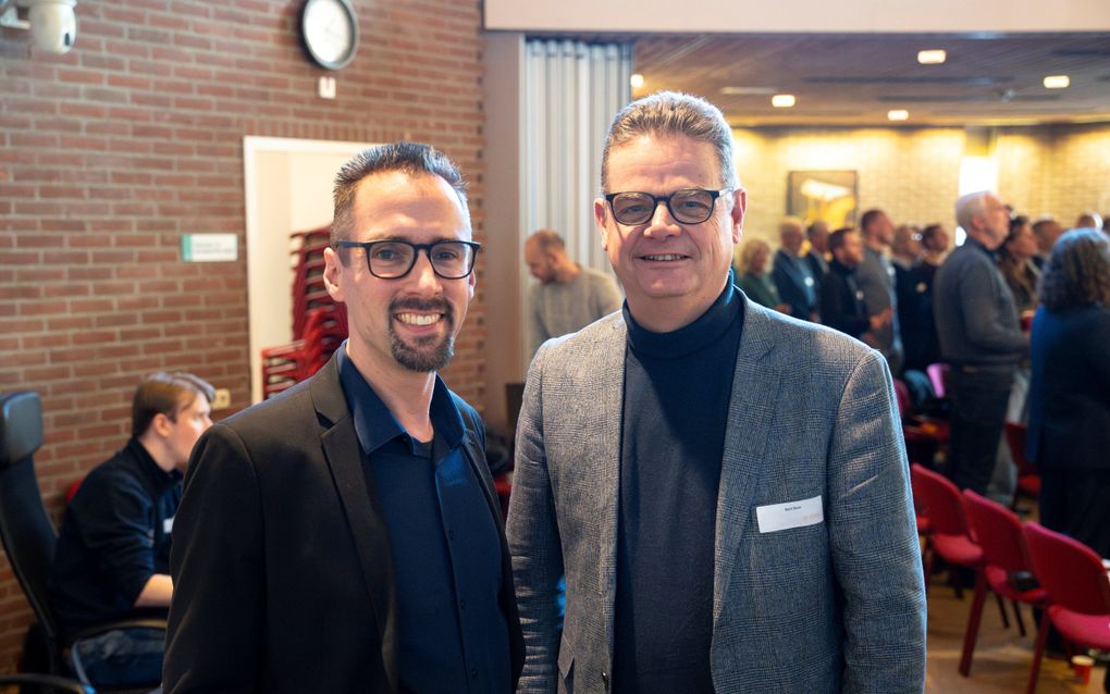 Twee mannen, beiden met een colbert en een bril, staan naast elkaar. Op de achtergrond staan andere mensen.