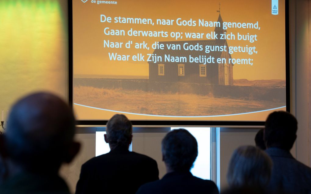 Beamerscherm met daarop de tekst van een berijmde psalm. Op de voorgrond de achterhoofden van enkele mensen die naar het scherm kijken.