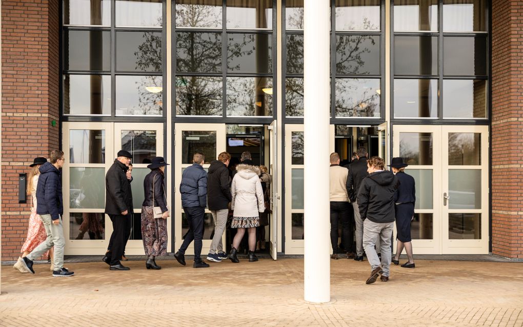 Mannen, vrouwen en jongeren lopen via verschillende deuren met veel glas een groot gebouw binnen.