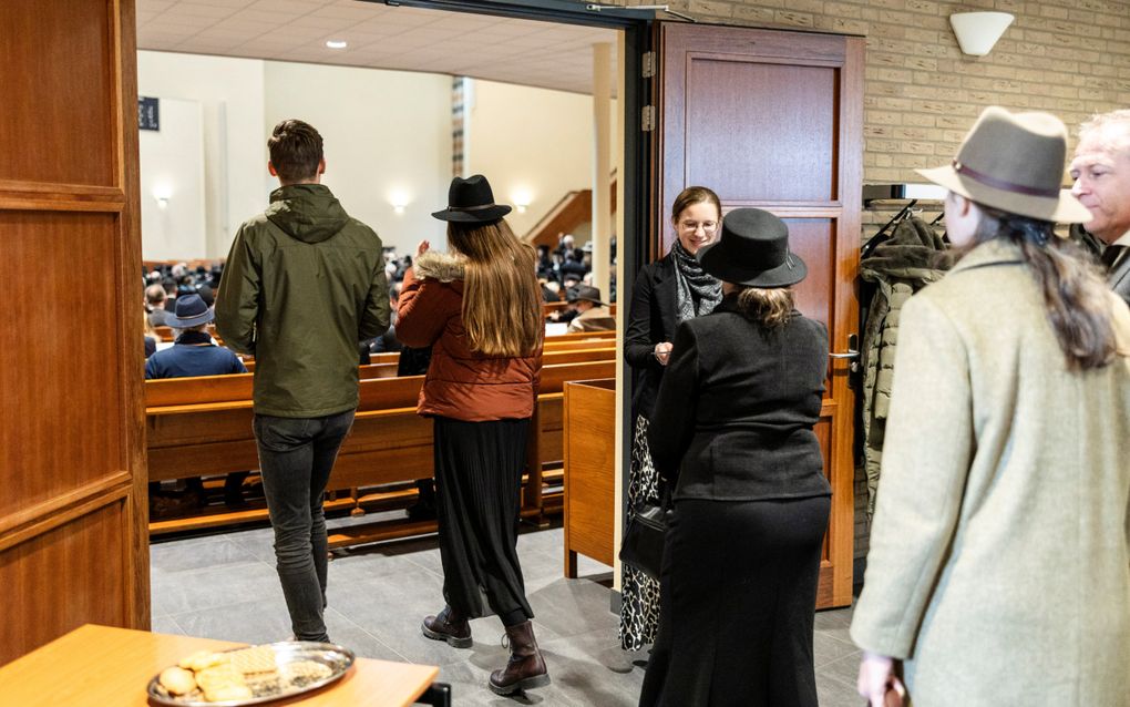 Een man en enkele vrouwen met hoed lopen een kerkzaal binnen waar al veel mensen zitten.
