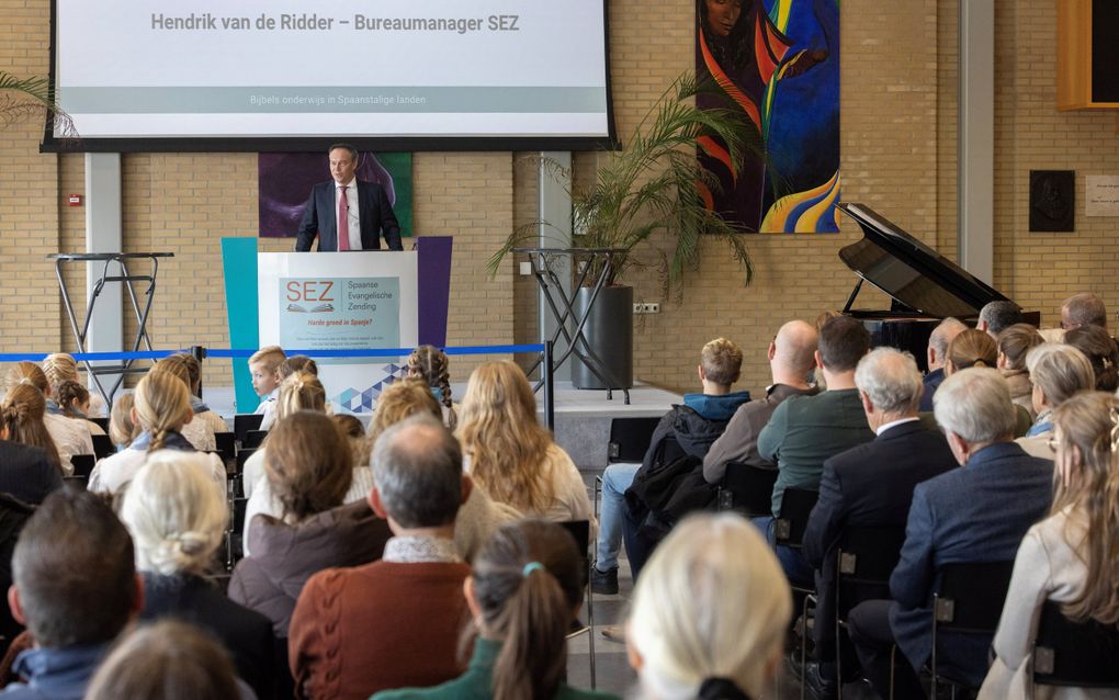Zaal vol mensen op de rug gezien. Voorin staat een man in een donker pak achter een katheder.
