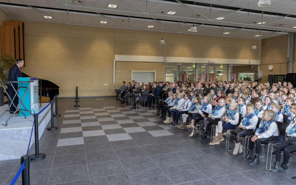 Zaal vol mensen, met voorin rijen kinderen. Voorin staat een man achter een katheder.