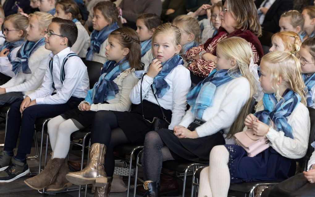Kinderen met witte blouses zitten op de voorste rij in een zaal met mensen.