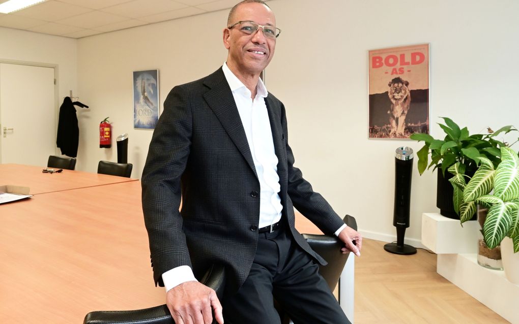 Een man met een bril, gekleed in een donker pak met een wit overhemd, zit op de rand van een grote tafel.