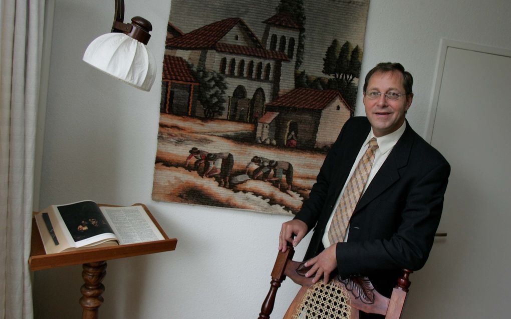 Man in donker pak met stropdas staat voor een wandkleed, leunend op een stoel.