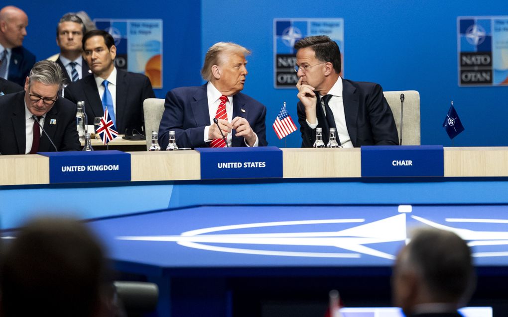 De Amerikaanse president met blauw jasje en roodwitte  stropdas. Naast hem NAVO-baas Mark Rutte, in donker gekleed. Op tafel staat tussen beide mannen in een Amerikaans vlaggetje. 