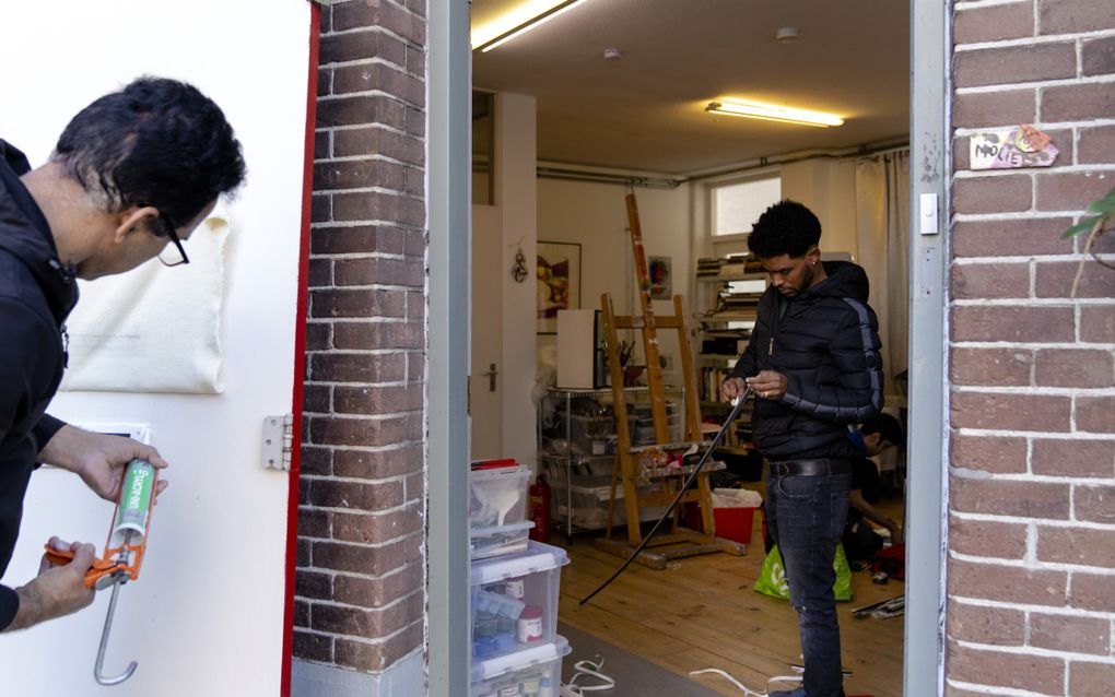 Man spuit kit rond een brievenbusopening in een deur; door de deuropening zie je een woonkamer waar iemand met een tochtstrip bezig is.