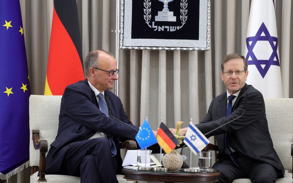 Twee mannen in pak zitten op witte stoelen aan een rond tafeltje, terwijl ze elkaars handen vastpakken. Op het tafeltje staan een kleine Europese, een Duitse en een Israëlische vlag; grote exemplaren daarvan staan achter de heren