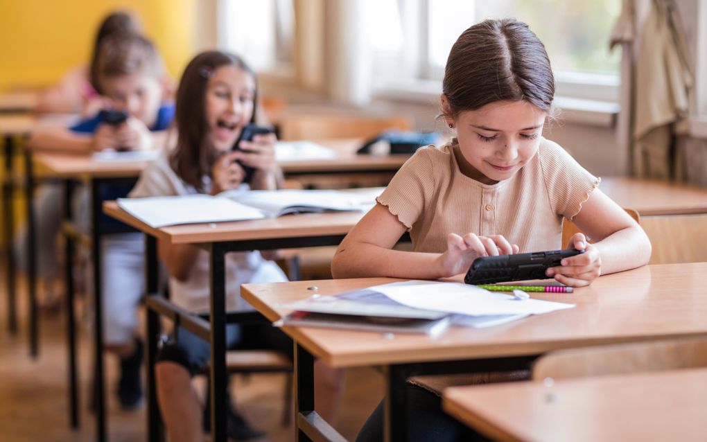 Meisje met beige shirt aan tafeltje in schoollokaal met smartphone in haar hand.