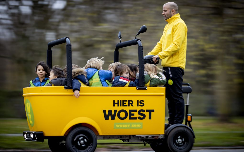 Een man met een geel hesje staat achterop een knalgele elektrische kar met daarin zo’n acht kinderen.