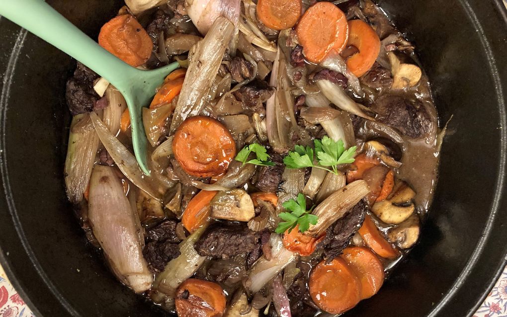 In een zwarte pan zit een mengsel van rundvlees, wortel, ui, champignons en groene kruiden in een saus.