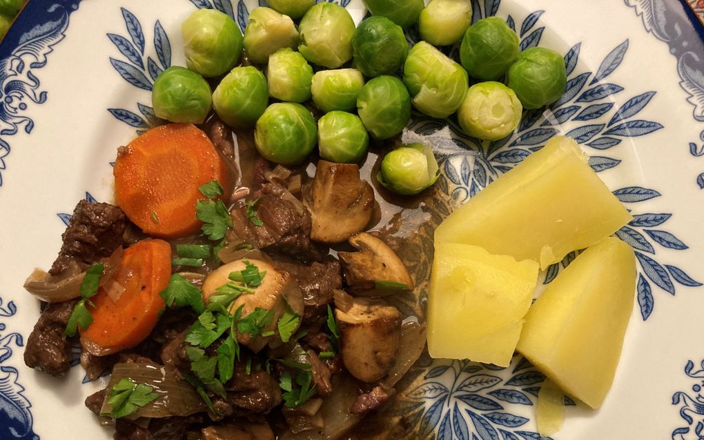 Op een crème bord met blauwe bloemen liggen stukjes rundvlees in een saus. Ernaast spruitjes, schijfjes wortel en stukjes aardappel.