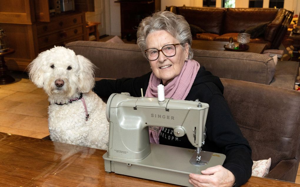 Oude dame achter een Singer naaimachine, met links van haar een witte hond.