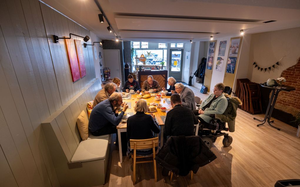 Tien mensen zitten aan een vierkante tafel in een inloophuis te eten.