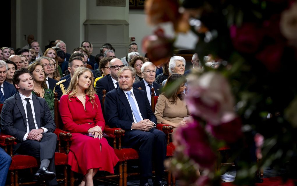 Vrouw in rode jurk zit op een stoel naast mannen in pak. De man rechts van haar kijkt opzij.