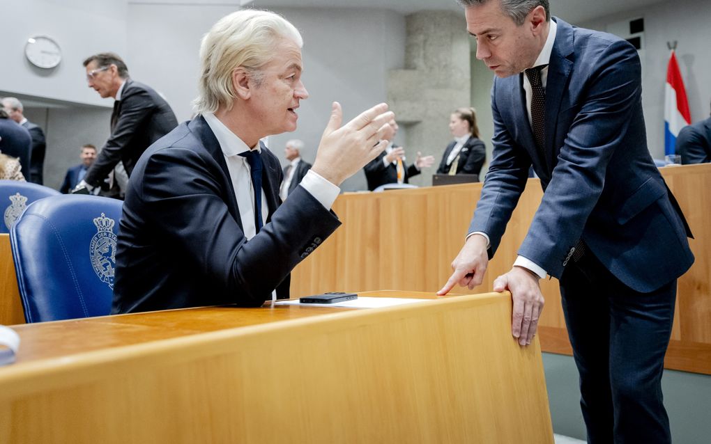 Man met witblond haar, zittend in de plenaire zaal van de Tweede Kamer, praat met man die bij hem staat.
