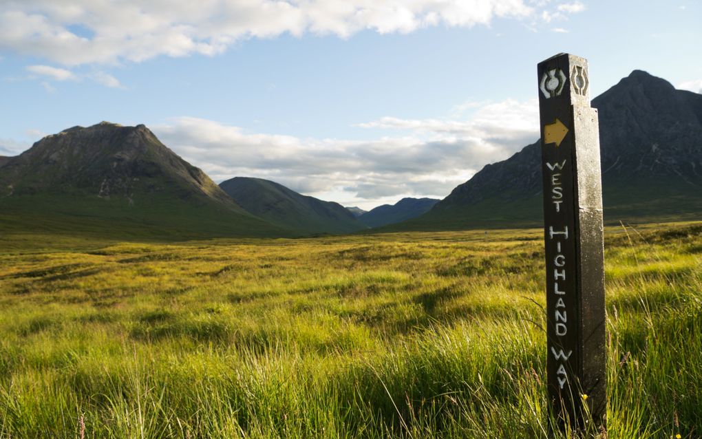 Groene grasvlakte in het zonlicht met bergen op de achtergrond. Rechts staat een paal met een wit teken en een gele pijl.