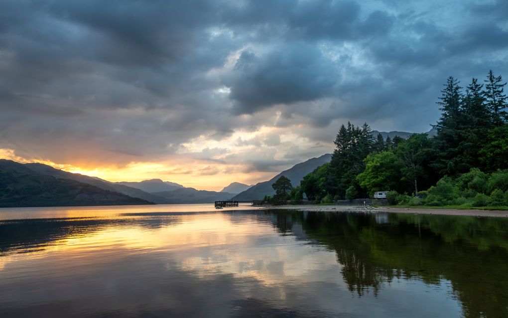 De zon verdwijnt achter bergen en weerspiegelt wolken in het water. Rechts is de oever bebost en ligt een steiger in het water.