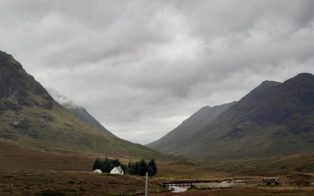 Grijsgroene rotsachtige bergen met een klein wit huisje en enkele naaldbomen op de voorgrond.