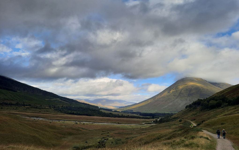 Een smal pad voert een vallei in met op de achtergrond een groene en steenachtige berg. Zonlicht schijnt op de berghelling. Op het pad lopen enkele wandelaars.