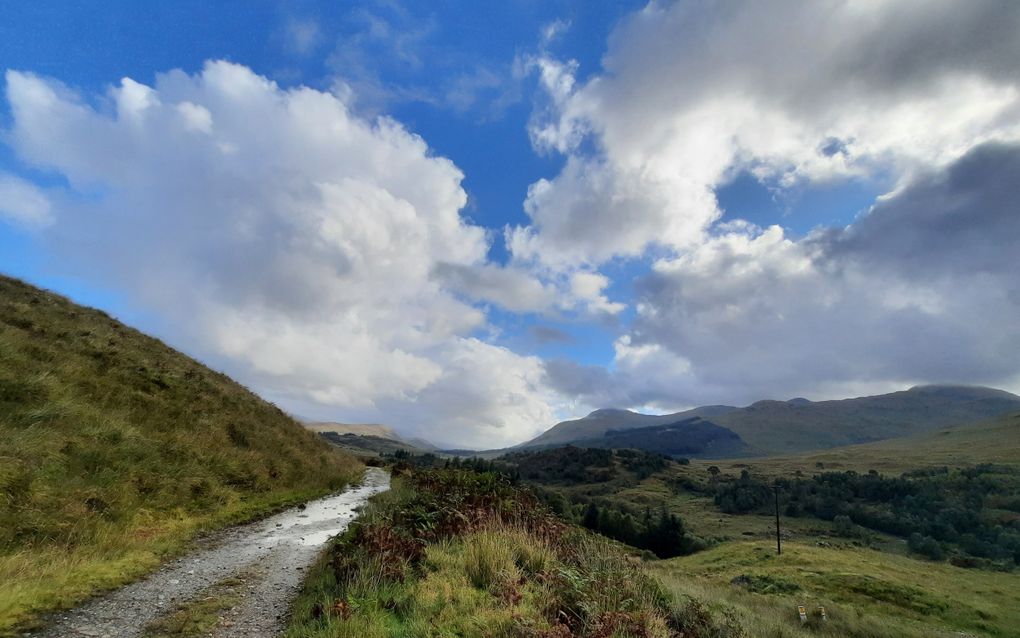 Een steenachtig pad maakt een flauwe bocht om een begroeide heuvel heen. In de verte is wolkenlucht en heuvelachtig grasland te zien.