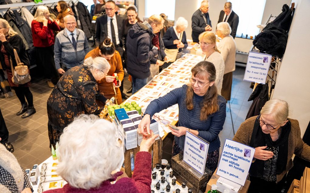 Vrouw geeft wisselgeld aan andere vrouw bij verkooptafel, waar papiertjes hangen met ‘’heerlijke zachte Israëlische douchegel”, ”Israëlische zeeppompjes’’ en dergelijke.